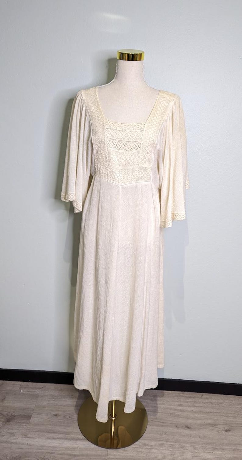 May include: A cream-colored vintage dress with a square neckline and bell sleeves. The bodice features a detailed lace panel. The dress is displayed on a mannequin with a gold base, set against a light blue wall and wooden floor.