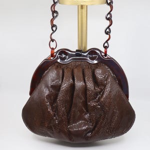 May include: A vintage brown handbag with a textured, snake-like pattern. The bag features a tortoiseshell-style frame and handle, and a decorative clasp. The handle is a chain of curved links. The bag is displayed on a gold stand.