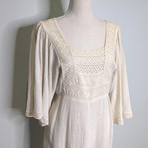May include: Cream-colored dress with a square neckline and flared sleeves. The bodice features intricate lace detailing, and the dress has a flowing, relaxed fit. The dress is displayed on a mannequin.