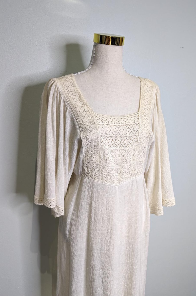 May include: Cream-colored dress with a square neckline and flared sleeves. The bodice features intricate lace detailing, and the dress has a flowing, relaxed fit. The dress is displayed on a mannequin.