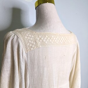 May include: Cream-colored dress with a square neckline and three-quarter sleeves. The neckline features a delicate, openwork lace detail. The dress is displayed on a mannequin, showcasing its textured fabric and design.