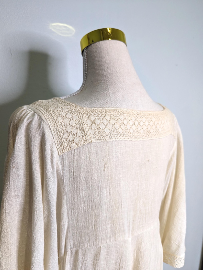 May include: Cream-colored dress with a square neckline and three-quarter sleeves. The neckline features a delicate, openwork lace detail. The dress is displayed on a mannequin, showcasing its textured fabric and design.