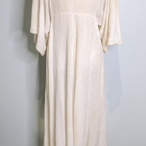 May include: Cream-colored dress with a square neckline and wide sleeves. The dress features a lace detail at the neckline and sleeve cuffs. The dress is displayed on a mannequin with a gold base.