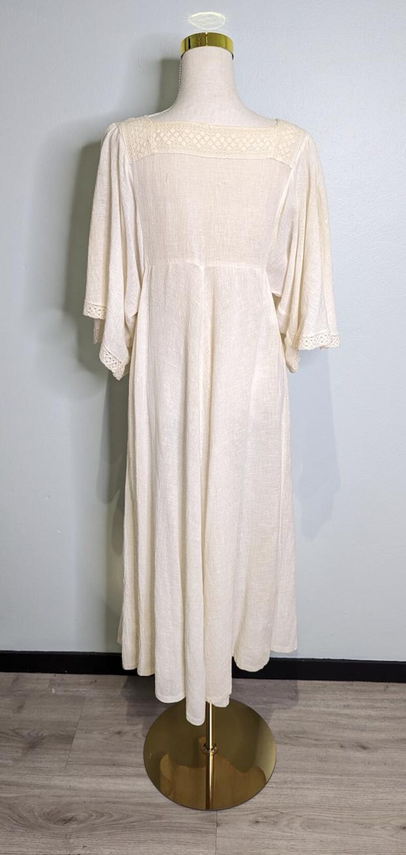 May include: Cream-colored dress with a square neckline and wide sleeves. The dress features a lace detail at the neckline and sleeve cuffs. The dress is displayed on a mannequin with a gold base.