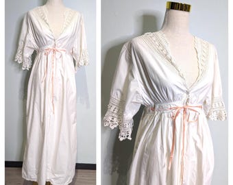 Puede incluir: Camisón blanco vintage con escote en V y ribete de encaje. El camisón presenta mangas cortas con puños de encaje y una cinta rosa atada a la cintura. Se exhibe en un maniquí.