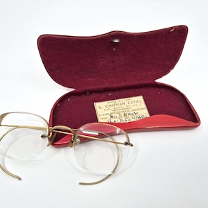 May include: Antique rimless spectacles with gold-coloured metal frames and clear lenses, next to an open red velvet-lined case. The case has a label with text. A classic eyewear accessory.
