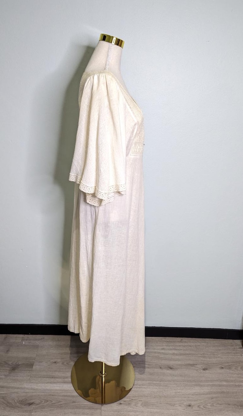 May include: Off-white vintage maxi dress with flared sleeves and a V-neckline. The dress features delicate lace trim on the sleeves and neckline. The dress is displayed on a gold dress form.