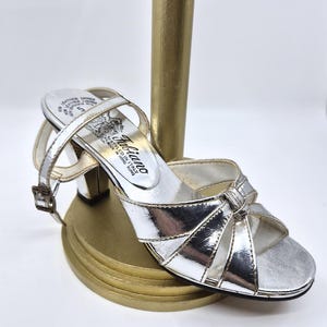 May include: Silver metallic sandal with a small heel and ankle strap. The sandal features a bow detail and is displayed on a gold-coloured stand. The brand name "Fabiano" is visible on the insole.