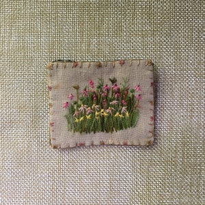 May include: A small embroidered patch featuring a cluster of pink and yellow flowers with green stems and leaves on a beige linen background.