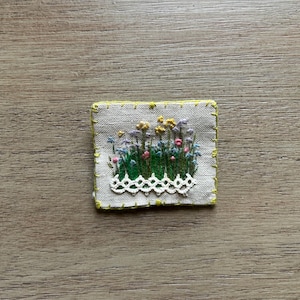 May include: A rectangular beige fabric patch featuring hand embroidery of green grass and small yellow, blue, and pink wildflowers. The patch is bordered by bright yellow stitching and includes a decorative white lace trim along the bottom edge.
