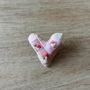 May include: A handmade heart-shaped fabric ornament. The ornament is made of pink and beige fabric patches, with embroidered red mushrooms and green leaves. The heart is placed on a wooden surface.