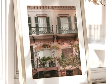 May include: A framed print of a building with balconies and shutters, displayed on a windowsill. The frame is a light color, and the image features a building with pink and peach tones. A vase of dried flowers sits nearby.
