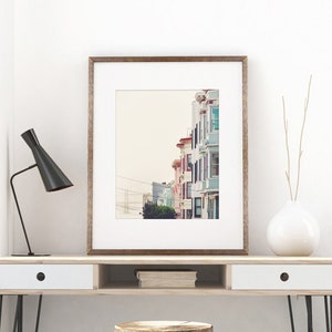 Photography print of pastel buildings in San Francisco with the Bay bridge in the bottom left background.  Colors include blue, red, pink, beige and green. Shown framed for reference only.