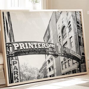 May include: Black and white photograph of a city street with a sign that reads "Printers Alley". The sign is an archway over the street, and there are buildings on either side of the street.