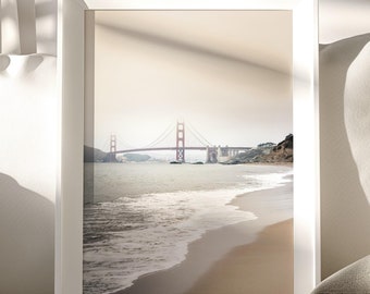 May include: A framed print of the Golden Gate Bridge in San Francisco. The image shows the bridge in the distance, with the beach and ocean in the foreground. The frame is white, and the overall color palette is neutral.