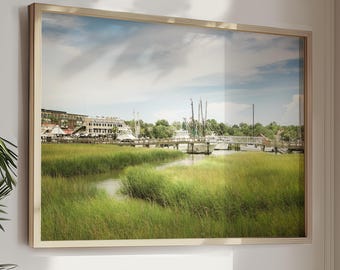 May include: A framed photograph of a waterfront scene with a wooden bridge, boats, and lush green marsh grasses. The sky is partly cloudy and the water is calm.