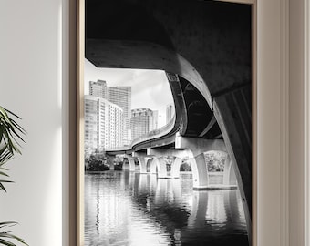 May include: A black and white framed wall art print featuring a low-angle perspective of a concrete bridge arching over a body of water, with city skyscrapers visible in the background under a bright sky, captured in a high-contrast monochromatic style.
