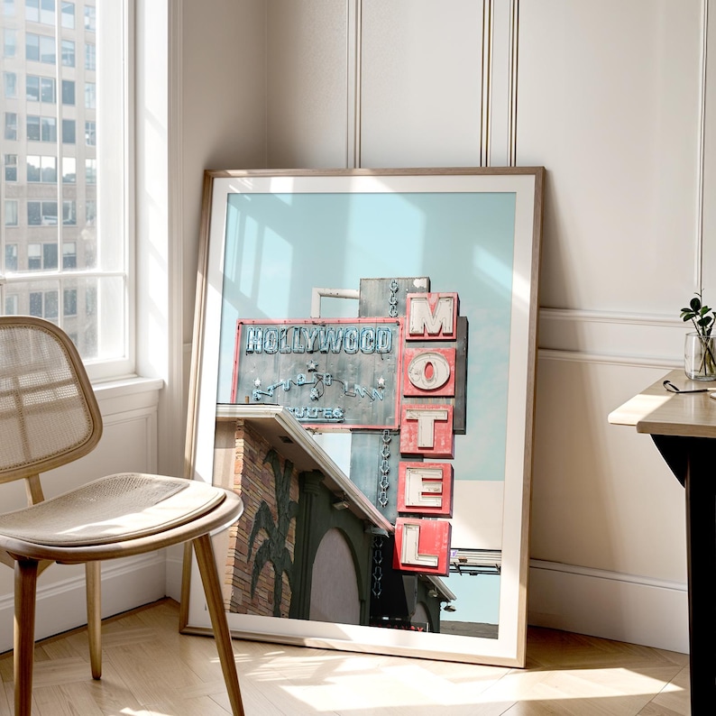May include: Framed print of the iconic Hollywood Motel sign. The sign features the words "Hollywood" and "Motel" in red and blue neon against a weathered metal background. The print is displayed in a light-colored wooden frame.
