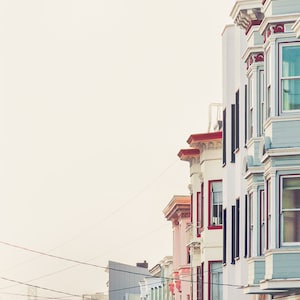 Photography print of pastel buildings in San Francisco with the Bay bridge in the bottom left background.  Colors include blue, red, pink, beige and green. Shown framed for reference only.