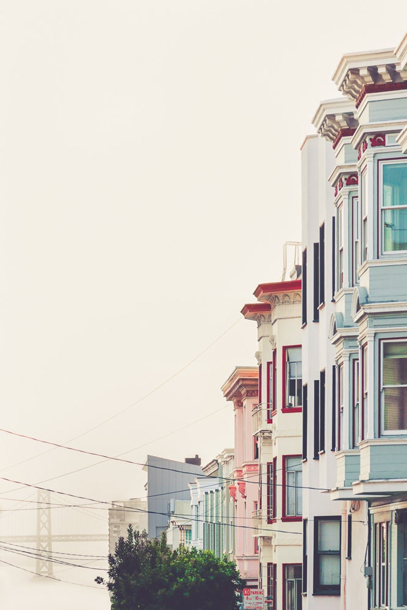 Photography print of pastel buildings in San Francisco with the Bay bridge in the bottom left background.  Colors include blue, red, pink, beige and green. Shown framed for reference only.