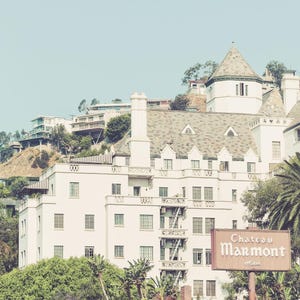 May include: A vintage photograph of the Chateau Marmont Hotel, a white building with a gray roof, set against a clear blue sky. A brown sign in front of the hotel reads "Chateau Marmont Hotel."