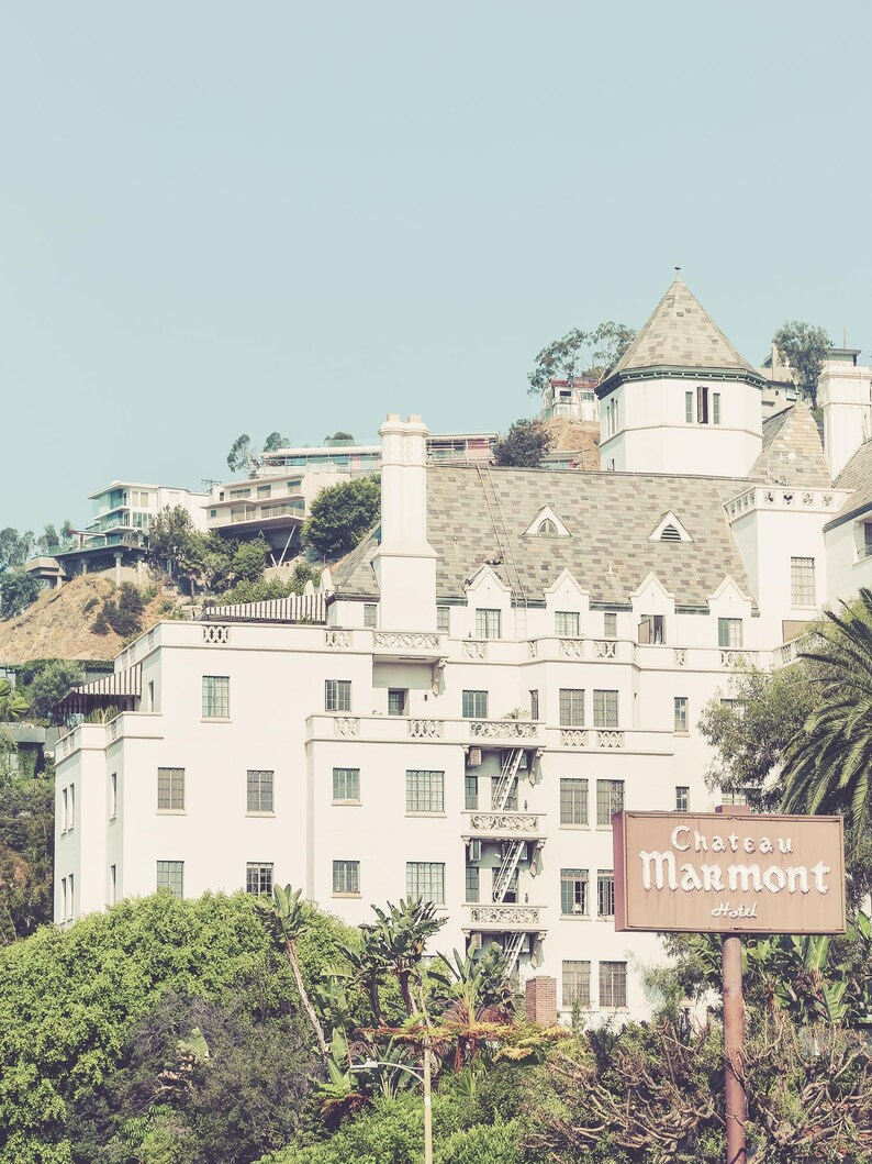 May include: A vintage photograph of the Chateau Marmont Hotel, a white building with a gray roof, set against a clear blue sky. A brown sign in front of the hotel reads "Chateau Marmont Hotel."
