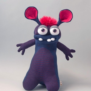 May include: A plush monster toy made of dark blue fabric with thin maroon horizontal stripes. It features large white eyes with purple pupils, a tuft of bright pink hair, large ears with pink inner lining, and two white teeth protruding from its mouth.