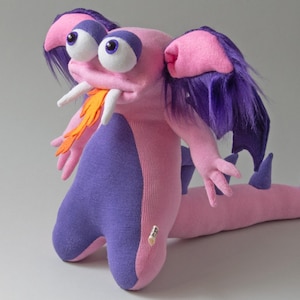 May include: A plush creature toy featuring a pink and purple body, large white eyes with purple lids, and purple hair. It has two white fangs, an orange felt flame protruding from its mouth, and a long tail with purple spikes along the back.
