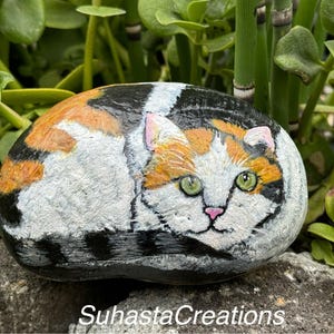 May include: Hand-painted rock art depicting a calico cat with green eyes, orange, black, and white fur. The rock is resting on a bed of green foliage.