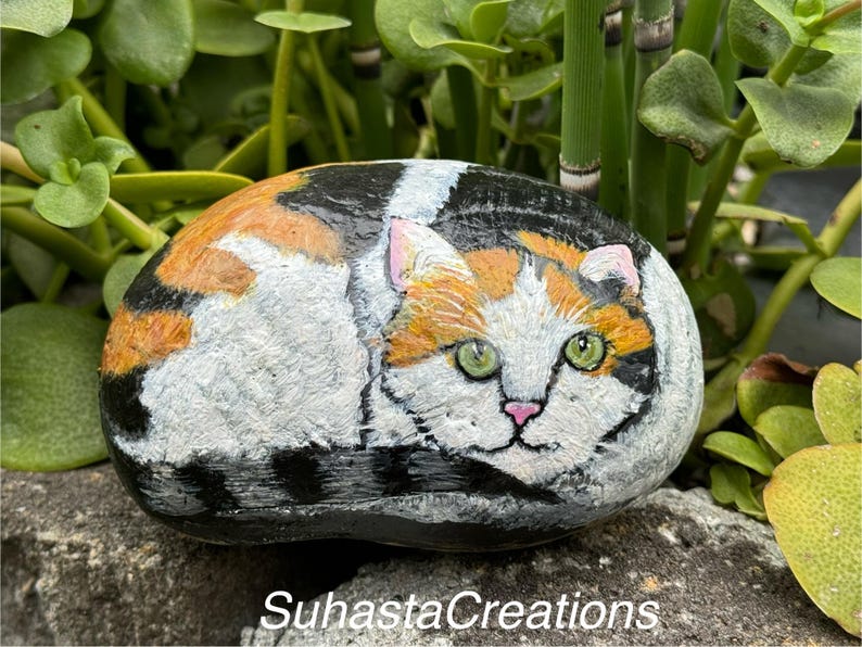 May include: Hand-painted rock art depicting a calico cat with green eyes, orange, black, and white fur. The rock is resting on a bed of green foliage.