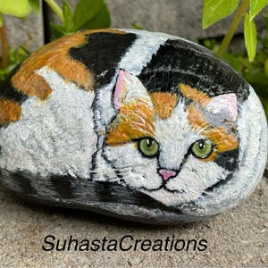 May include: Hand-painted rock art depicting a calico cat with green eyes, orange, black, and white fur. The cat is curled up in a sleeping position.