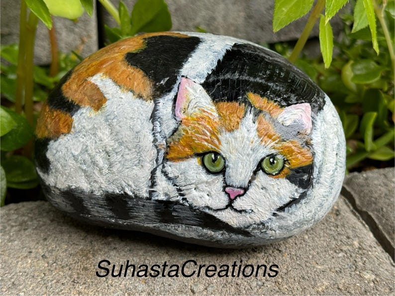May include: Hand-painted rock art depicting a calico cat with green eyes, orange, black, and white fur. The cat is curled up in a sleeping position.