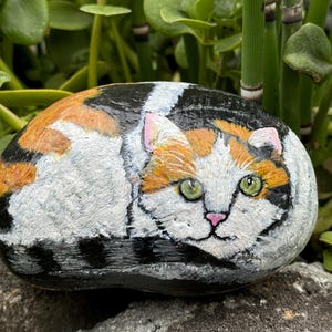 May include: Hand-painted rock art depicting a calico cat with orange, white, and black markings. The cat is painted in a realistic style with green eyes and a pink nose.