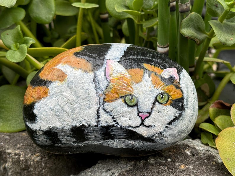 May include: Hand-painted rock art depicting a calico cat with orange, white, and black markings. The cat is painted in a realistic style with green eyes and a pink nose.
