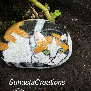 May include: Hand-painted rock art depicting a calico cat with green eyes. The cat is painted in a realistic style and is curled up on a rock. The rock is painted to look like a natural stone.