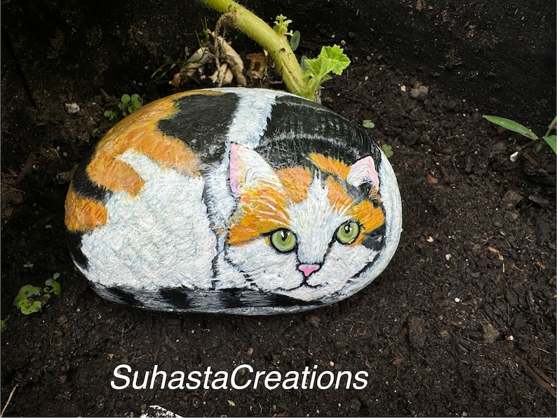 May include: Hand-painted rock art depicting a calico cat with green eyes. The cat is painted in a realistic style and is curled up on a rock. The rock is painted to look like a natural stone.