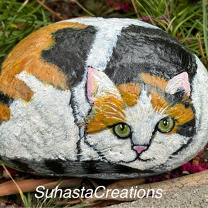 May include: Hand-painted rock art depicting a calico cat with green eyes, orange, black, and white fur. The cat is curled up in a sleeping position.