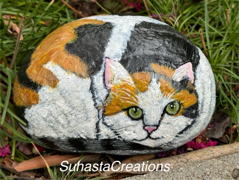 May include: Hand-painted rock art depicting a calico cat with green eyes, orange, black, and white fur. The cat is curled up in a sleeping position.