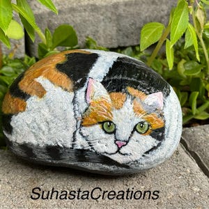 May include: Hand-painted rock art depicting a calico cat with green eyes, curled up in a sleeping position. The rock is painted in shades of white, orange, black, and brown.