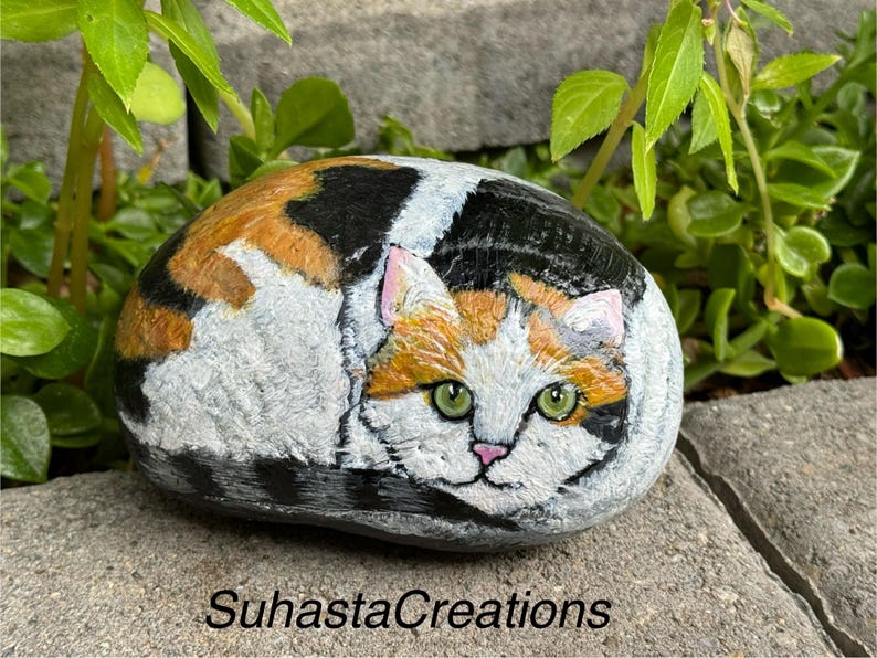 May include: Hand-painted rock art depicting a calico cat with green eyes, curled up in a sleeping position. The rock is painted in shades of white, orange, black, and brown.