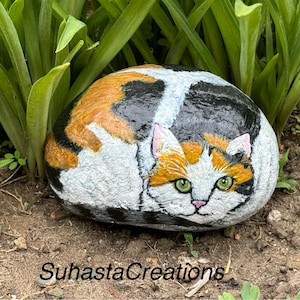 May include: A hand-painted rock depicting a calico cat curled up, with green eyes and orange, black, and white fur. The rock is sitting on a bed of brown dirt and green grass.