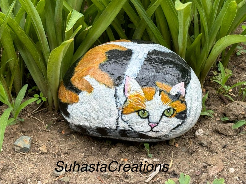 May include: A hand-painted rock depicting a calico cat curled up, with green eyes and orange, black, and white fur. The rock is sitting on a bed of brown dirt and green grass.