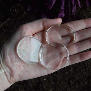 Op de afbeelding: Drie heldere, ronde kristallen stenen worden in een hand gehouden. De stenen zijn transparant met kleine onvolkomenheden. De achtergrond is een donker, getextureerd oppervlak met hints van paars en bruin.