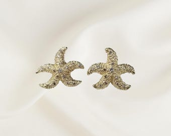 May include: A pair of gold-colored starfish earrings. Each earring has five points and is encrusted with small, sparkling stones. The earrings are set against a soft, off-white background.