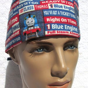 May include: A blue and red scrub hat with a Thomas the Tank Engine design. The hat features the text "Really Reliable Thomas", "Right On Time", "Ready Set Go", "1st Class Thomas", "1 Blue Engine", "You've Got A Ticket To Ride", "Right On Time", "1 Blue Engine", and "Full Steam Ahead".