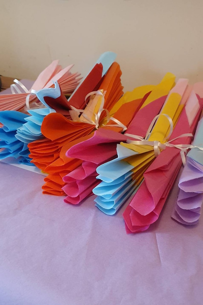 May include: A stack of colorful tissue paper fans tied with white ribbon. The fans are in shades of pink, orange, yellow, blue, and purple.