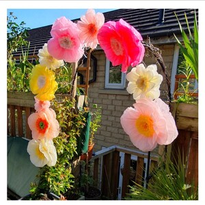 May include: A decorative archway adorned with large, multi-coloured paper flowers in shades of bright pink, soft peach, pale yellow, and cream. The flowers are arranged along a curved frame, featuring layered petals and distinct centres.