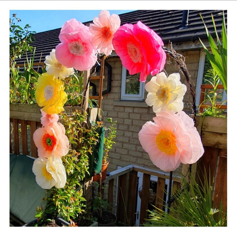 May include: A decorative archway adorned with large, multi-coloured paper flowers in shades of bright pink, soft peach, pale yellow, and cream. The flowers are arranged along a curved frame, featuring layered petals and distinct centres.