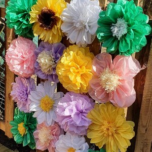 May include: A wall decorated with tissue paper flowers in various colors, including green, white, yellow, pink, and purple. The flowers are arranged in a random pattern and are attached to a wooden fence.