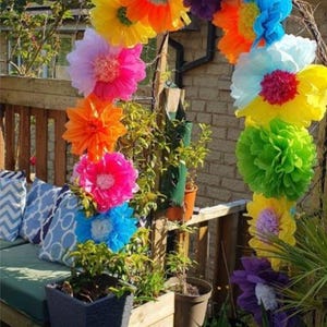 May include: A decorative archway constructed from large, multi-coloured tissue paper flowers in shades of yellow, orange, pink, purple, blue, and green, arranged in a curved formation and positioned outdoors on a wooden deck.